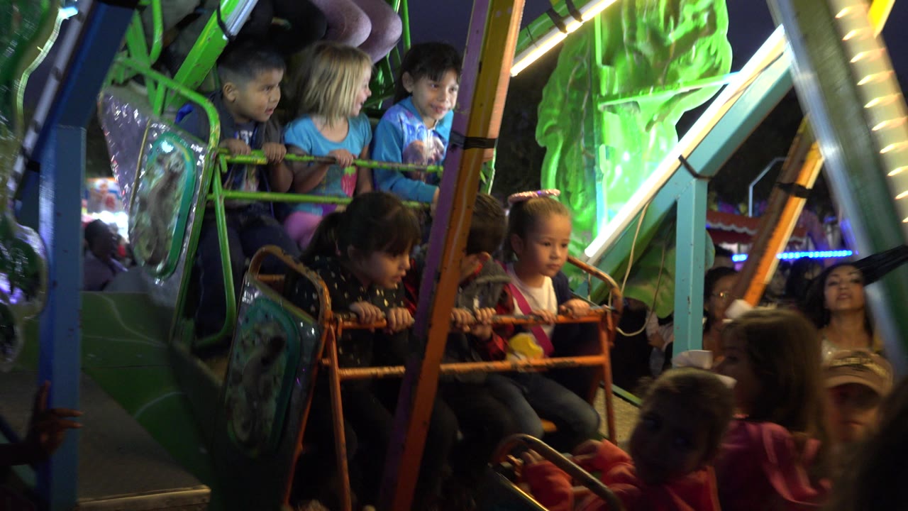 Children sit behind orange safety bars on a nighttime amusement ride in San Jose Del Cabo, Mexico, with green panels and bright ride lights around them.