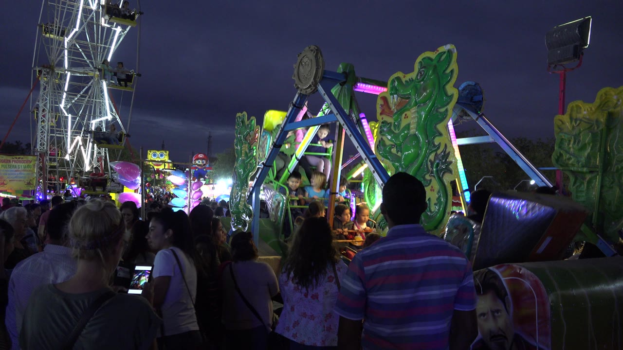 Spectators fill the foreground as a green and blue carnival ride moves through the midground in San Jose Del Cabo, Mexico, with bright white ride lights behind it.