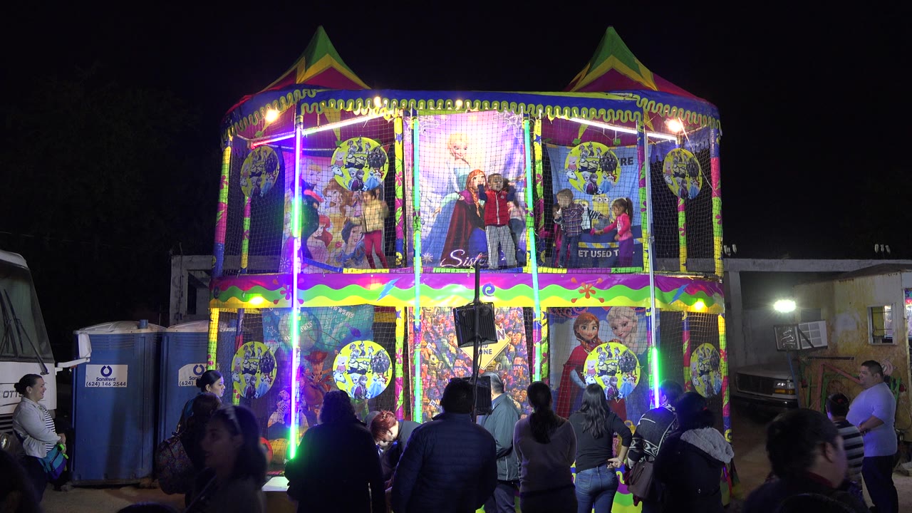 The lit children’s show structure remains centered in San Jose Del Cabo, Mexico, while adults and children shift position near the camera under strong colored LEDs.