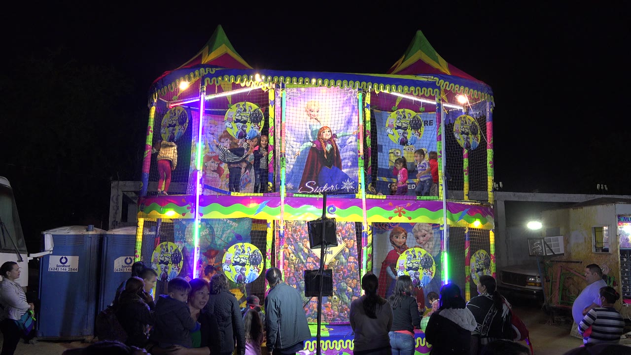 A neon-lit inflatable play structure in San Jose Del Cabo, Mexico shows children behind mesh panels, adults clustered below, and portable toilets to the side.