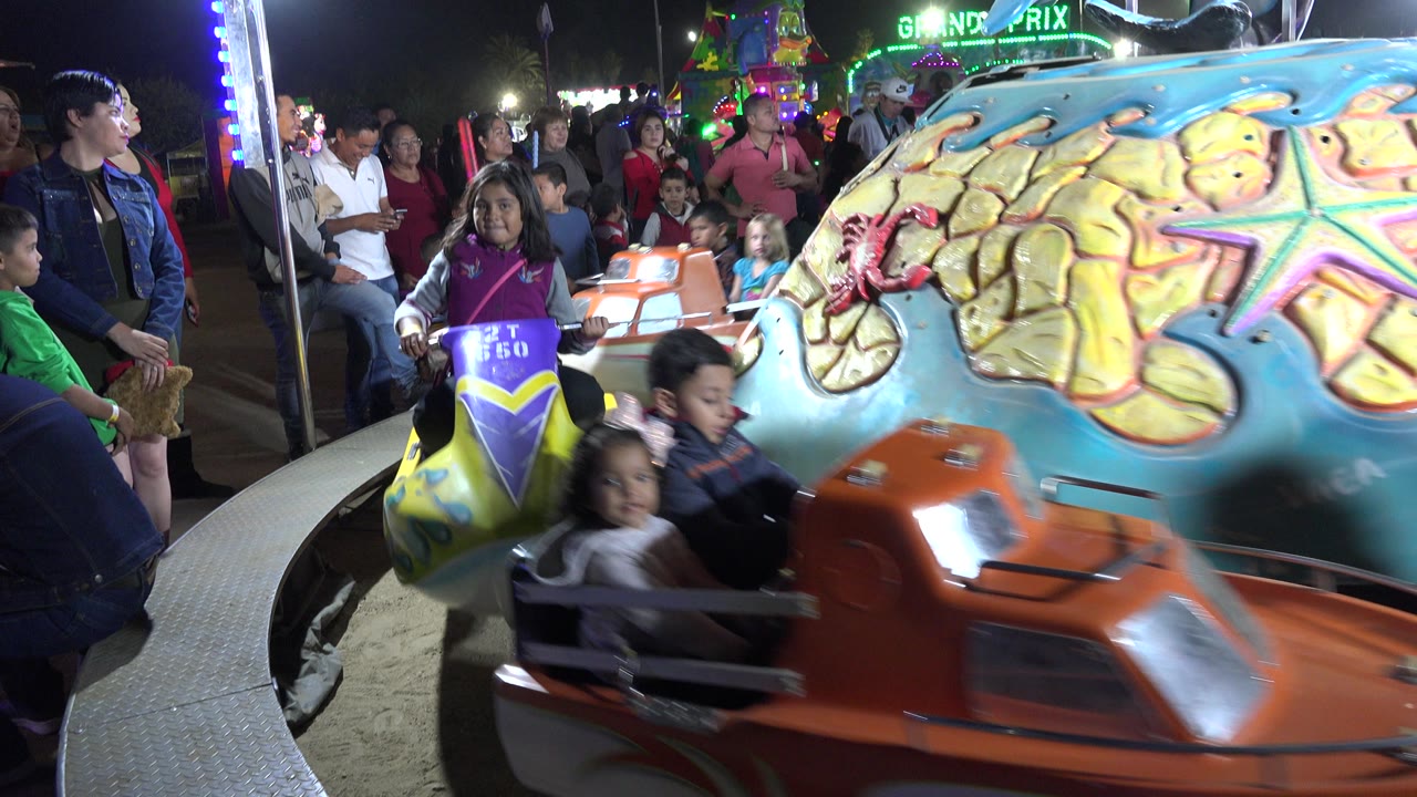At 0:01 in San Jose del Cabo, Mexico, an orange boat seat blurs across the foreground while a girl in a purple seat and spectators remain visible behind it.