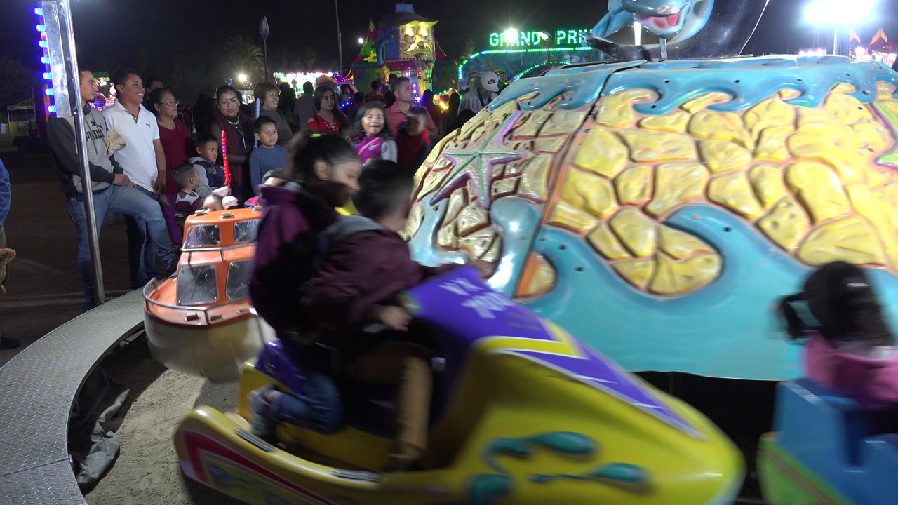 In San Jose del Cabo, Mexico, a yellow ride car with children moves across the foreground beside a painted center structure while families watch from the background.