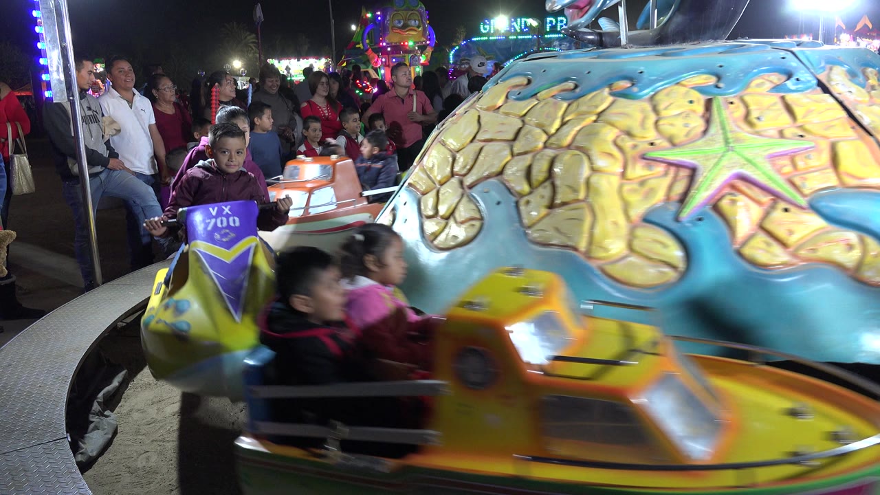 Children ride colorful carousel cars as spectators watch behind them.