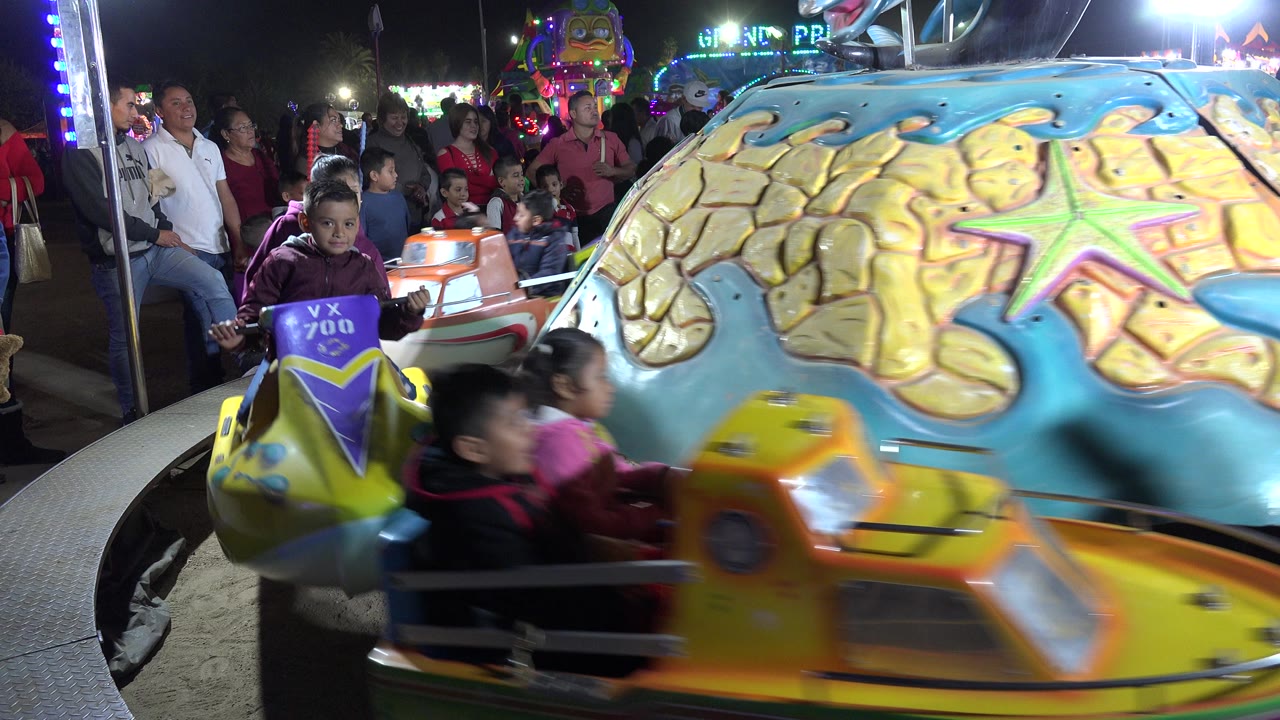 A child in a purple carousel car looks toward the camera as yellow and orange ride cars move through the foreground in San Jose del Cabo, Mexico.