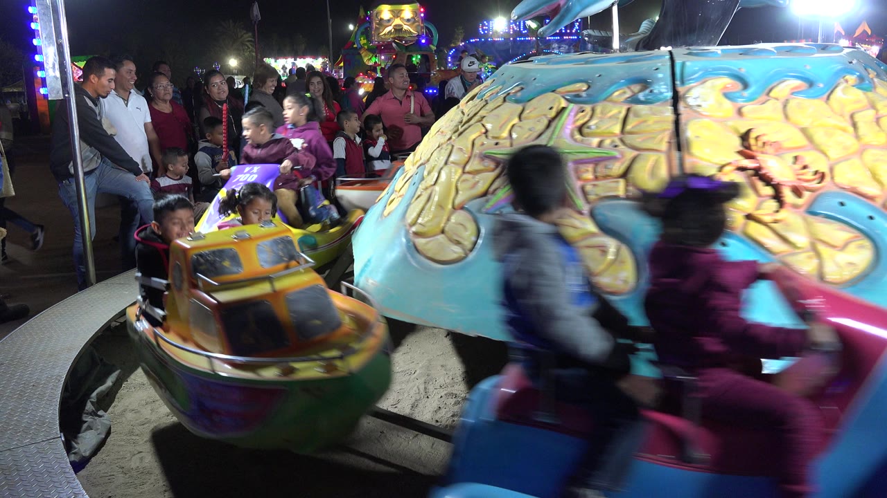 A yellow carousel car approaches the foreground while blurred children ride past the painted central structure and families stand in the background in San Jose Del Cabo, Mexico.