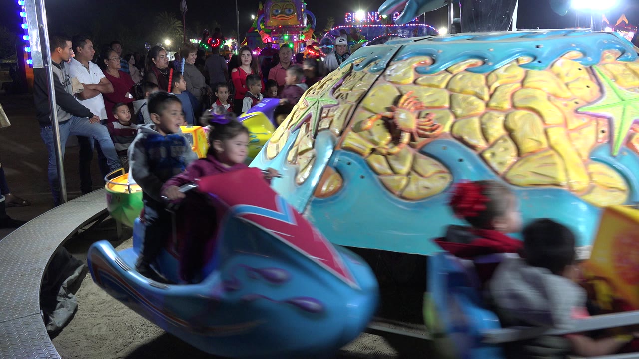A blue jet ski shaped car carrying children moves across the foreground in San Jose del Cabo, with the sea themed ride center and waiting crowd behind it.