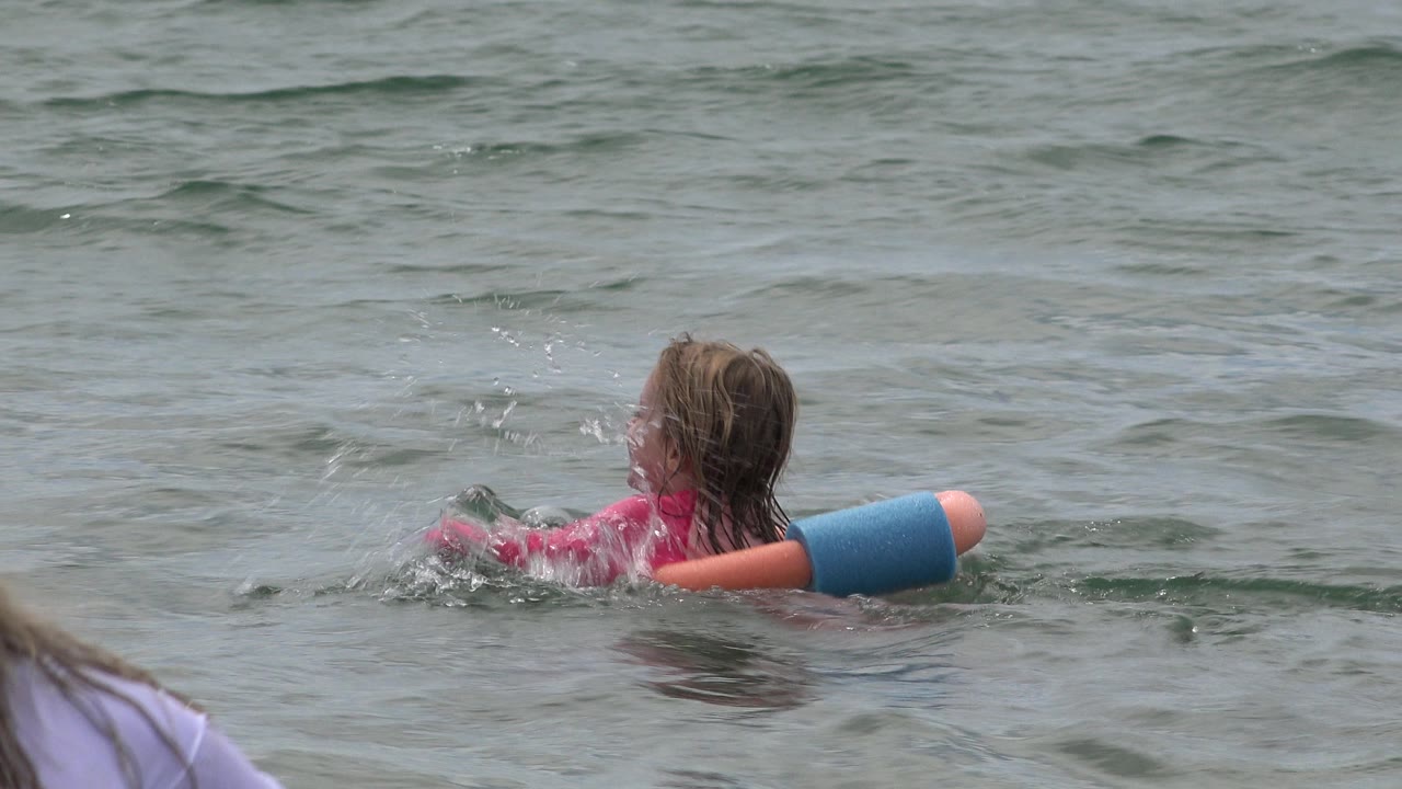 The girl continues paddling in San Jose Del Cabo ocean water, with splashes around her arms and the blue pool float visible at her side.