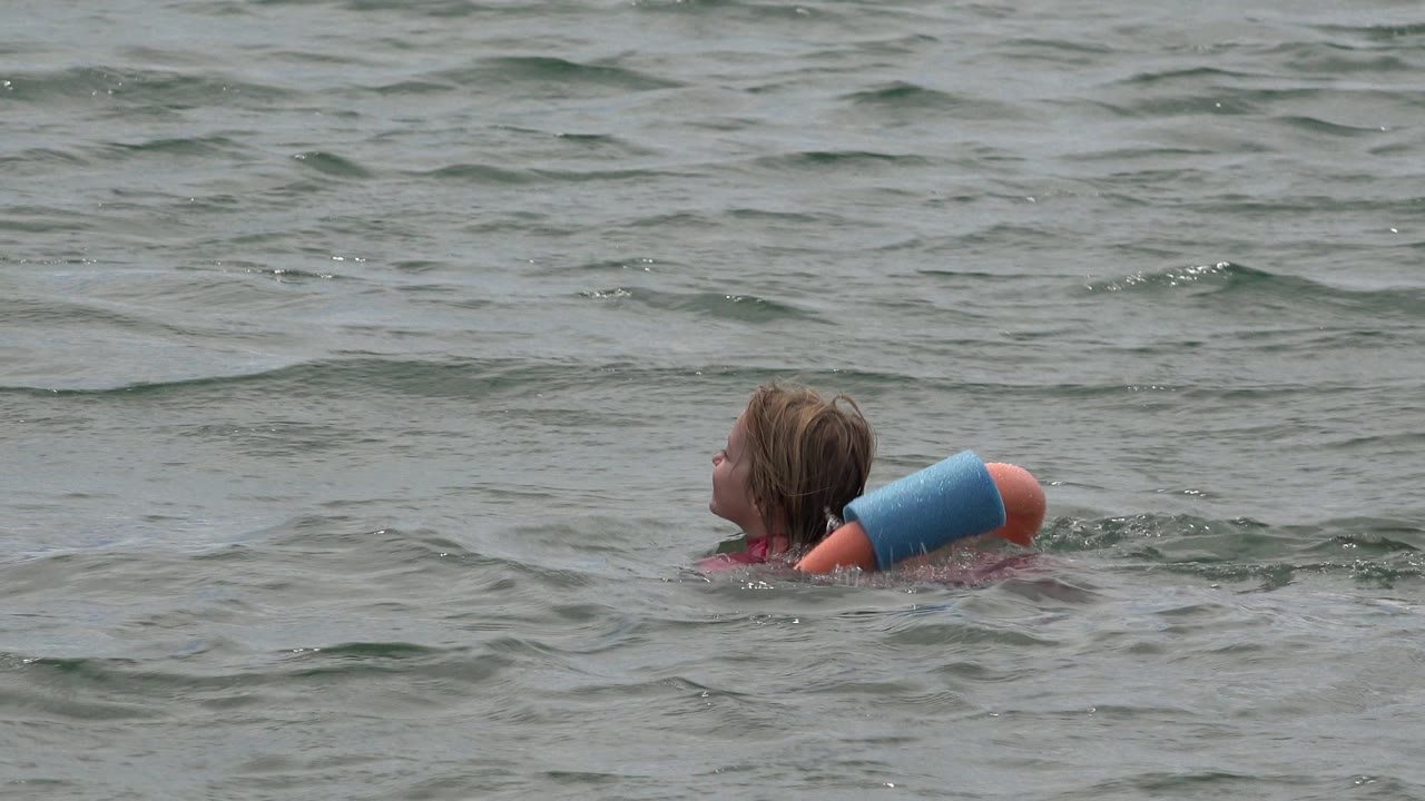 A young girl in San Jose del Cabo floats leftward in green-gray open water with a blue and orange swim ring supporting her upper body.