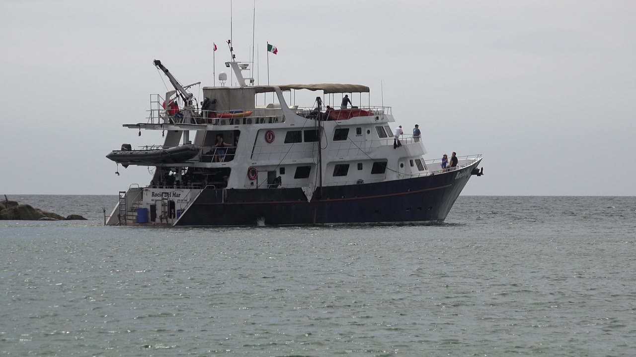 Tour-style passenger boat on open water in San Jose Del Cabo, Mexico, with small distant people at the bow and deck, rocks to the left, and a pale gray sky over the horizon.