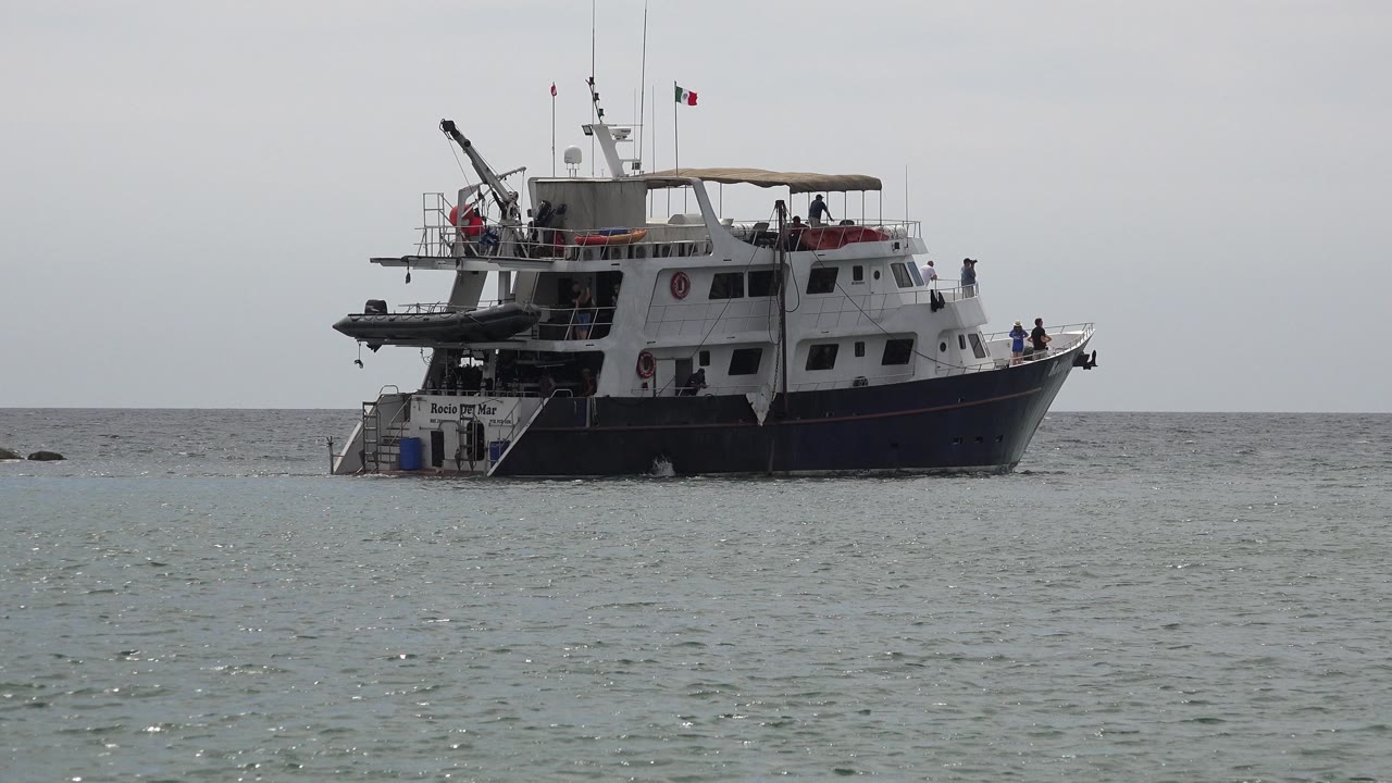 The boat remains centered offshore in San Jose del Cabo, Mexico, with small people visible near the deck and bow and a flat horizon behind it.