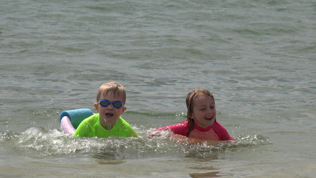 Two children face the camera while floating and splashing in shallow beach water.