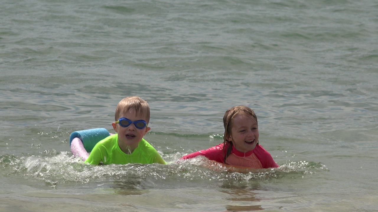 The boy and girl remain side by side in San Jose del Cabo, Mexico, with small waves and splashes around their foam pool noodles.