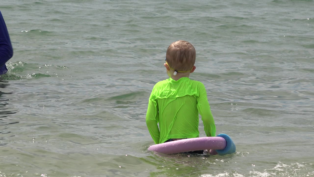 A boy in a bright green rash guard faces away in shallow water in San Jose del Cabo, Mexico, with purple and blue foam flotation noodles around his waist.