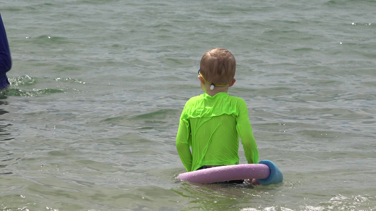 The boy remains centered in the San Jose del Cabo water, facing away as small ripples move around the purple and blue flotation noodles.
