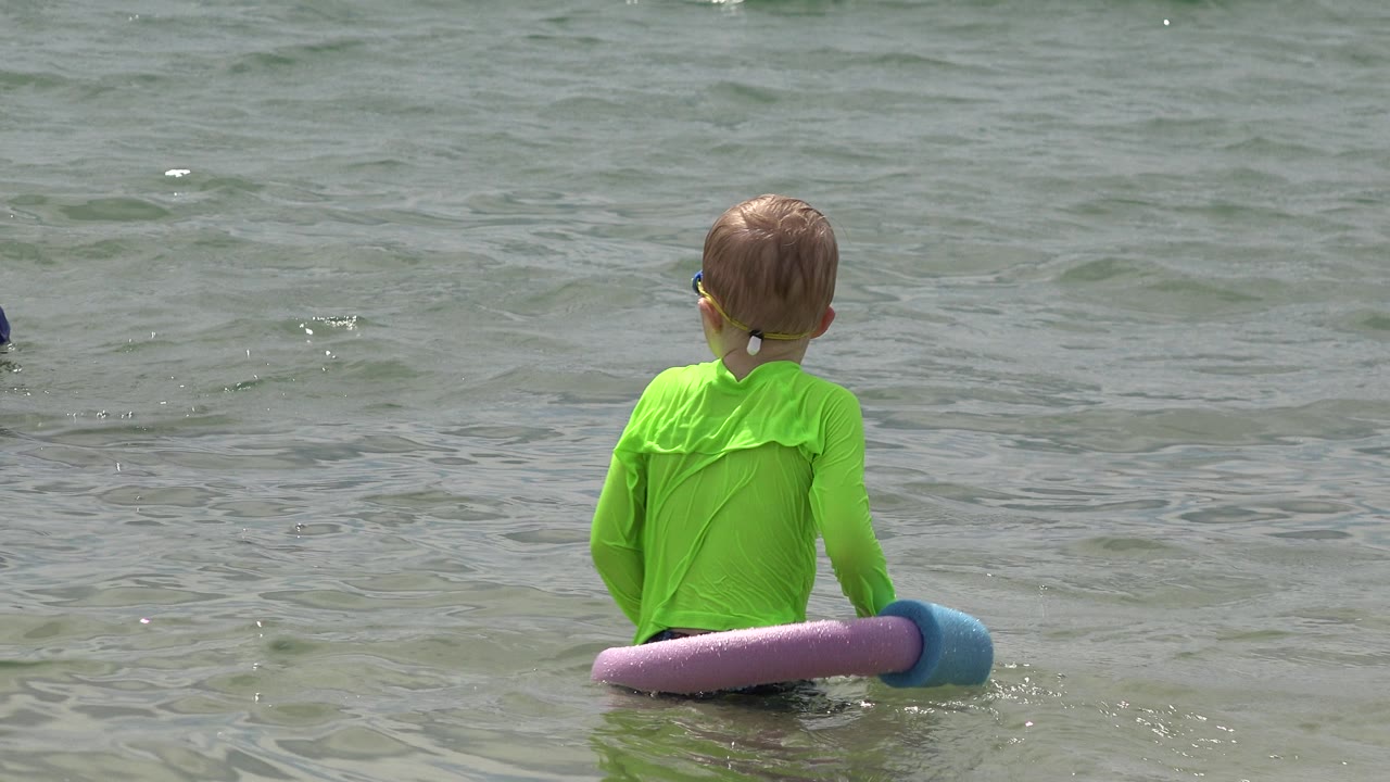 Boy in green sits in shallow lake water with foam flotation noodles.