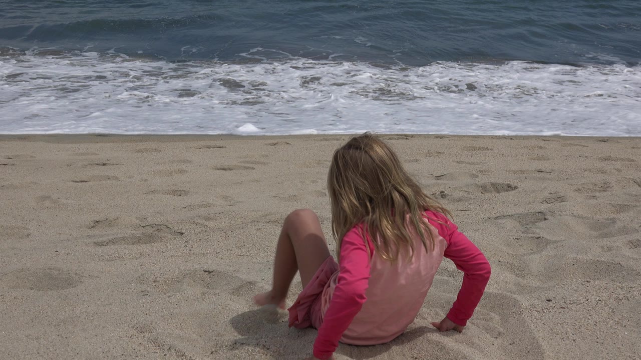 Child seated on sand facing ocean waves.