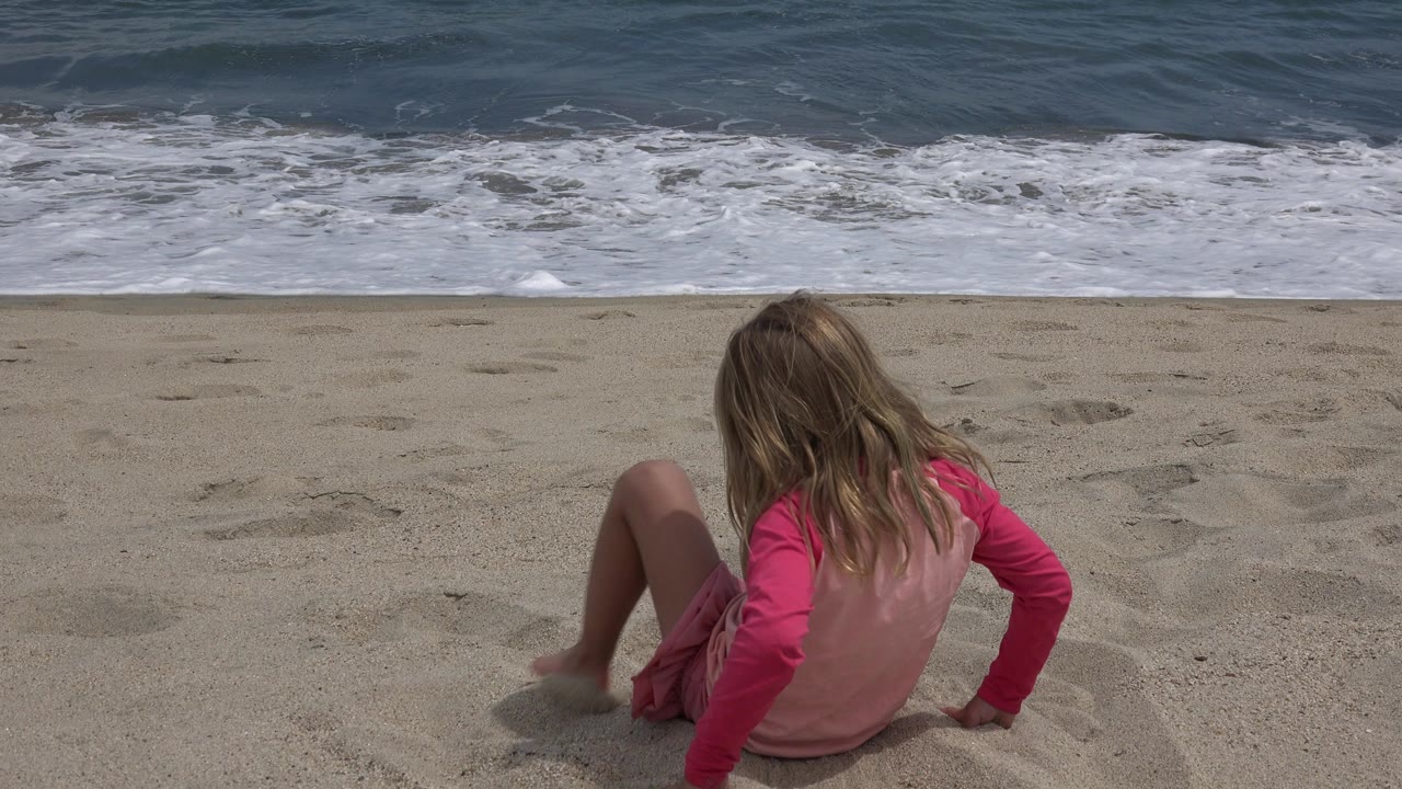 The child remains seated on the sand in San Jose Del Cabo, Mexico, with one hand braced behind them and foamy water rolling near the shoreline.