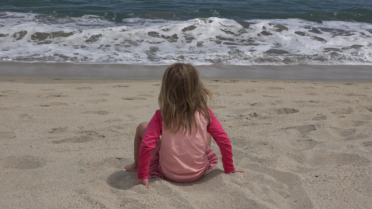 Blond child seated on sand facing ocean waves.