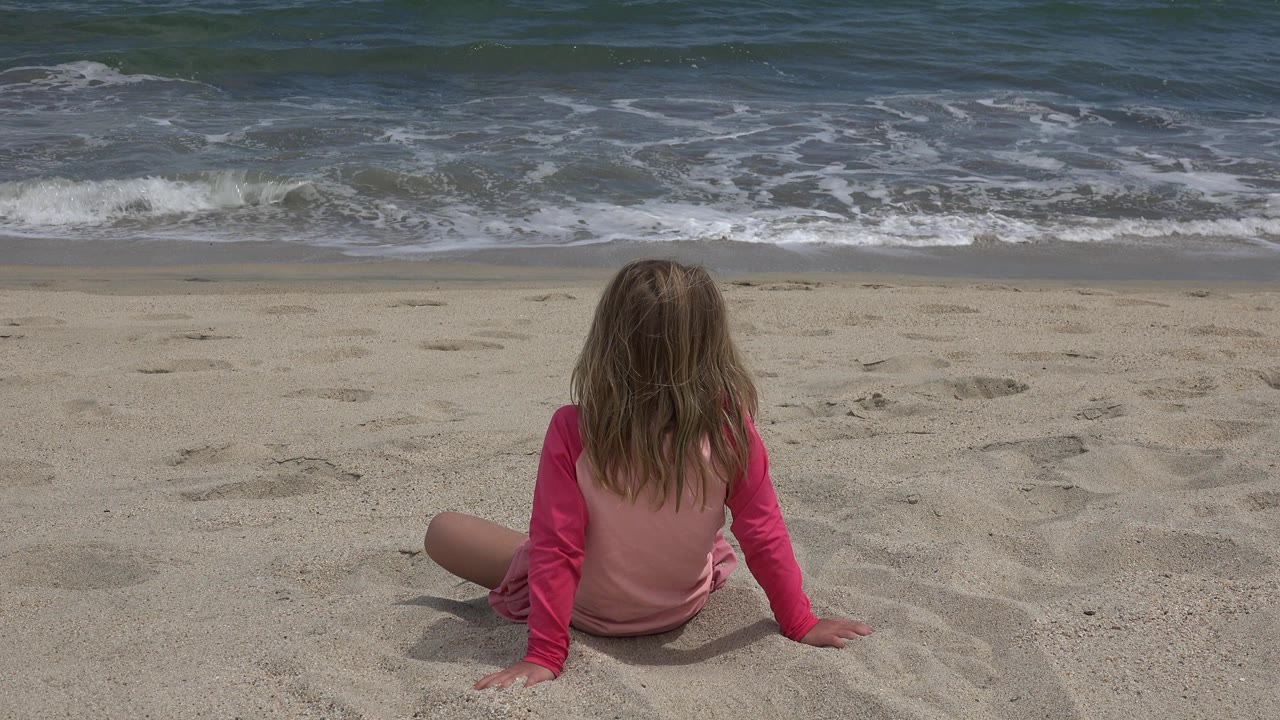 At 0:00, a young girl in a pink top sits with her back to camera on a San Jose del Cabo beach as white surf breaks across the upper frame.