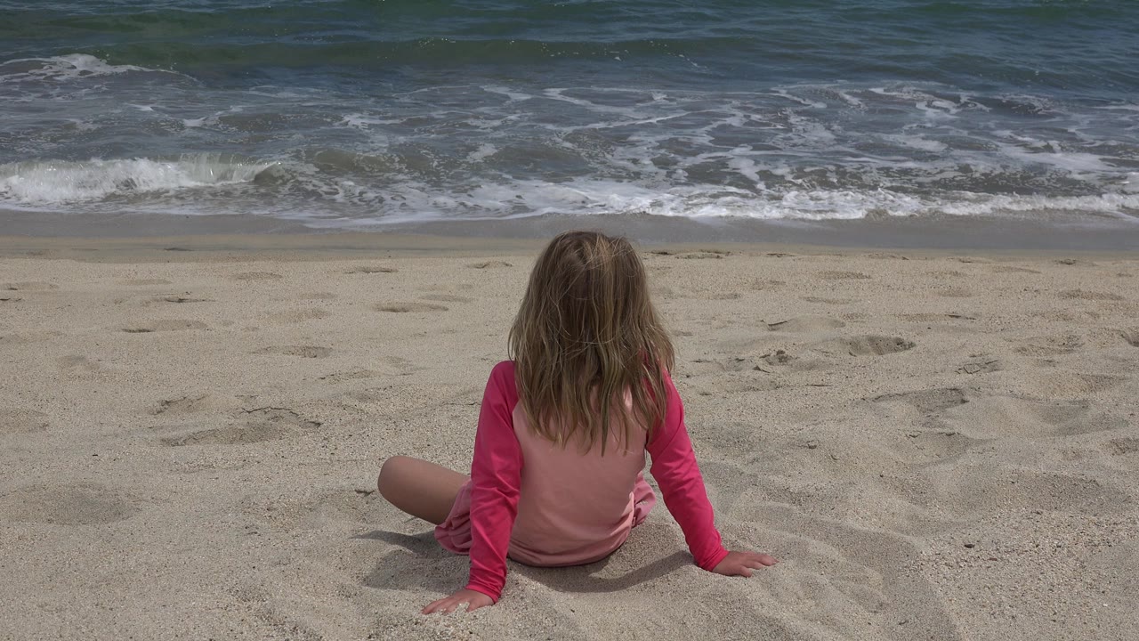 At 0:01, the seated girl remains centered low in frame while darker water and a thin line of foam move behind her on the San Jose del Cabo shoreline.