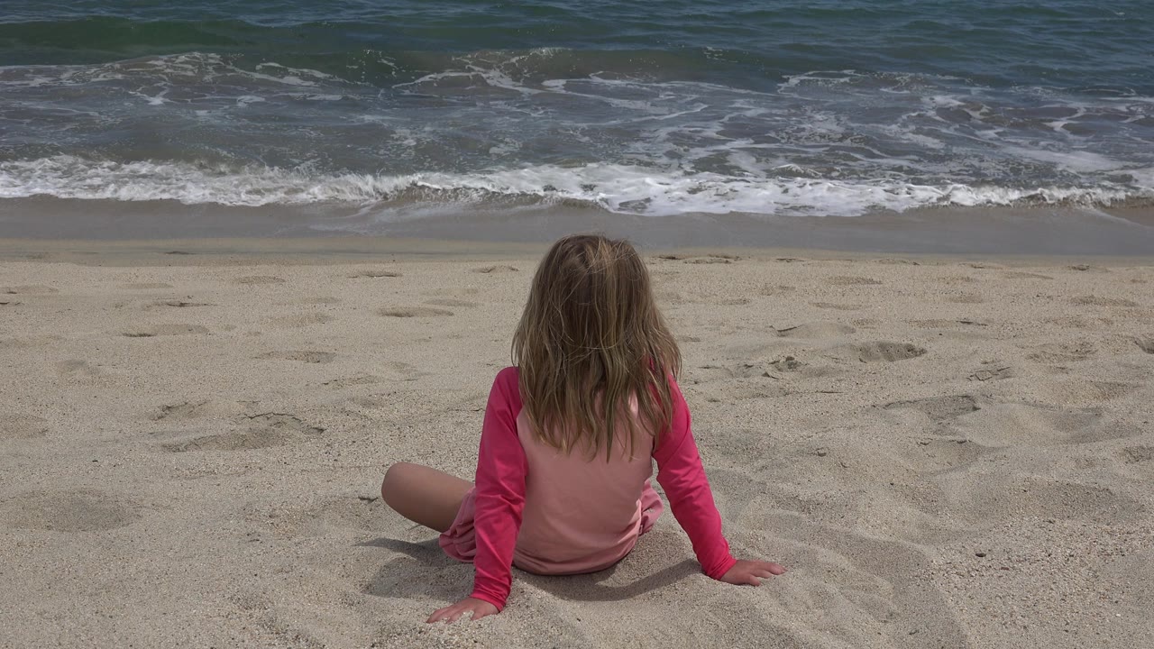 Child in pink sitting before ocean surf.