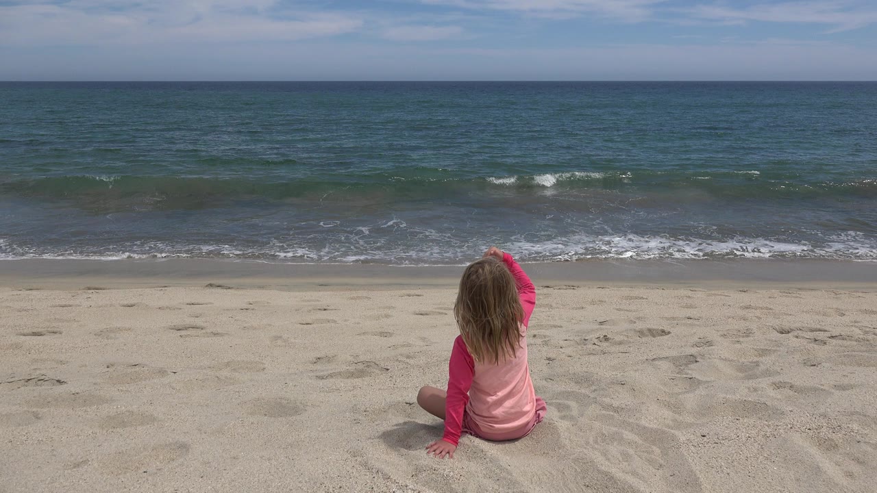 Child sitting on sand facing ocean waves.