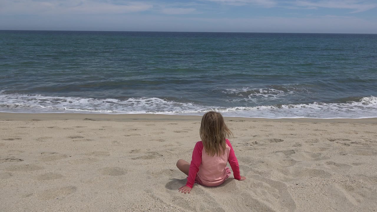 Blonde child in pink sitting on sand facing the ocean.