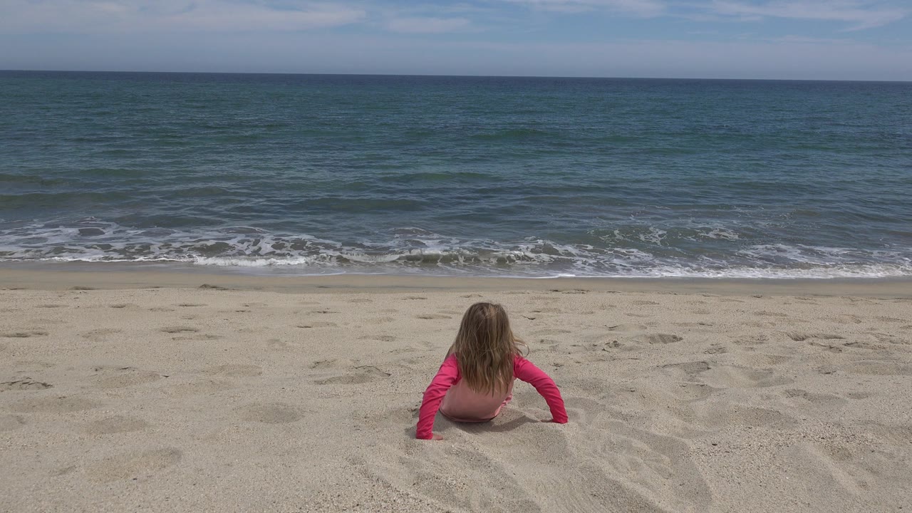 Small child sitting on sand facing ocean surf.