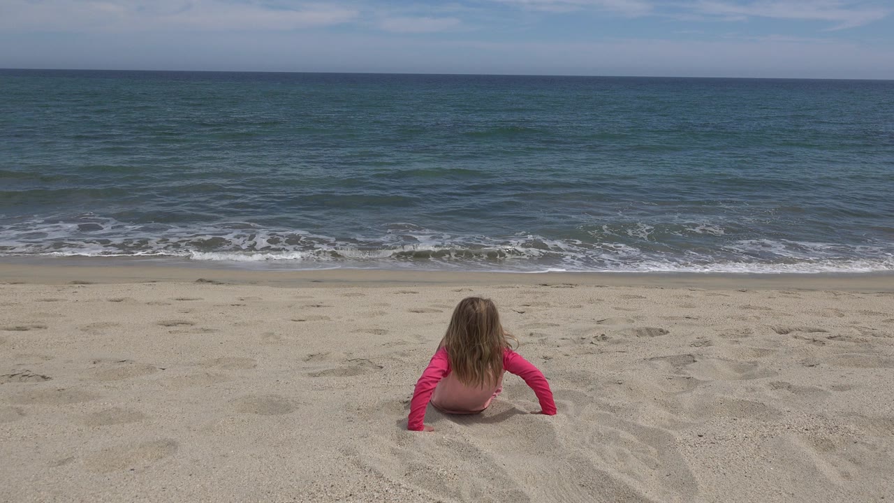 The child remains seated with arms braced in the sand, looking toward a band of foamy surf and blue-green water at San Jose del Cabo.