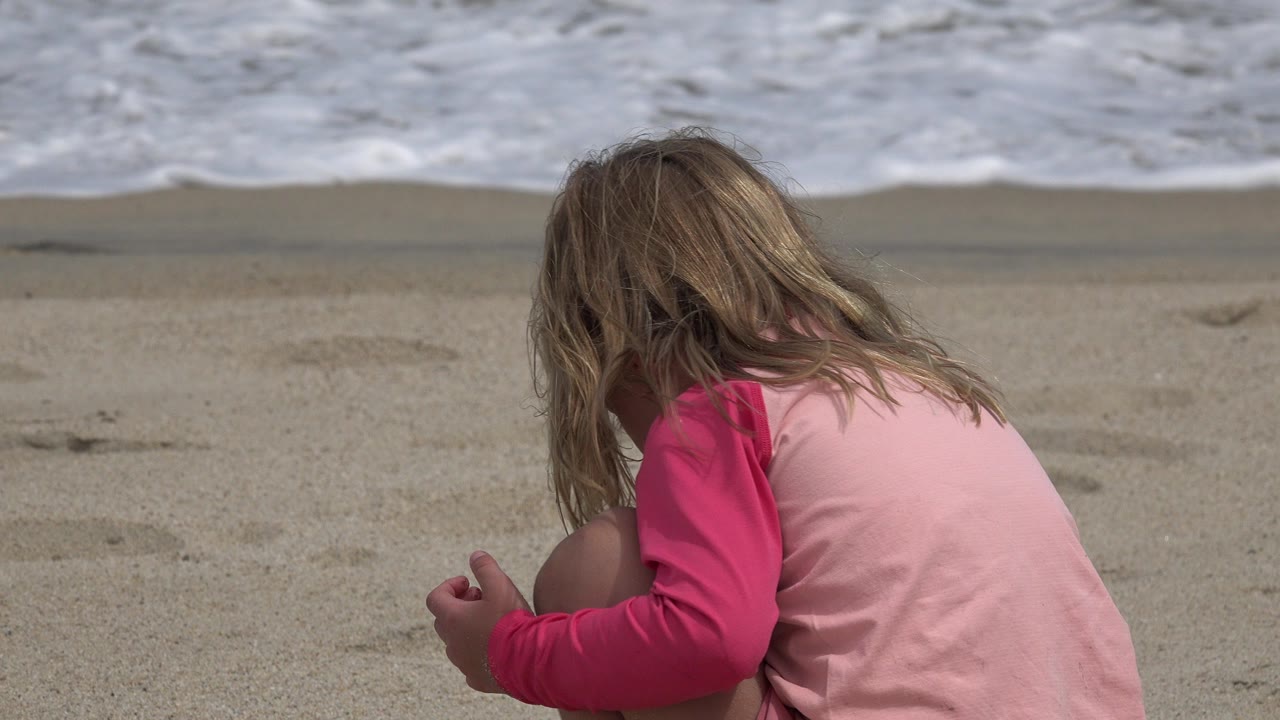 Blonde child in pink crouching on sand with ocean waves behind her.