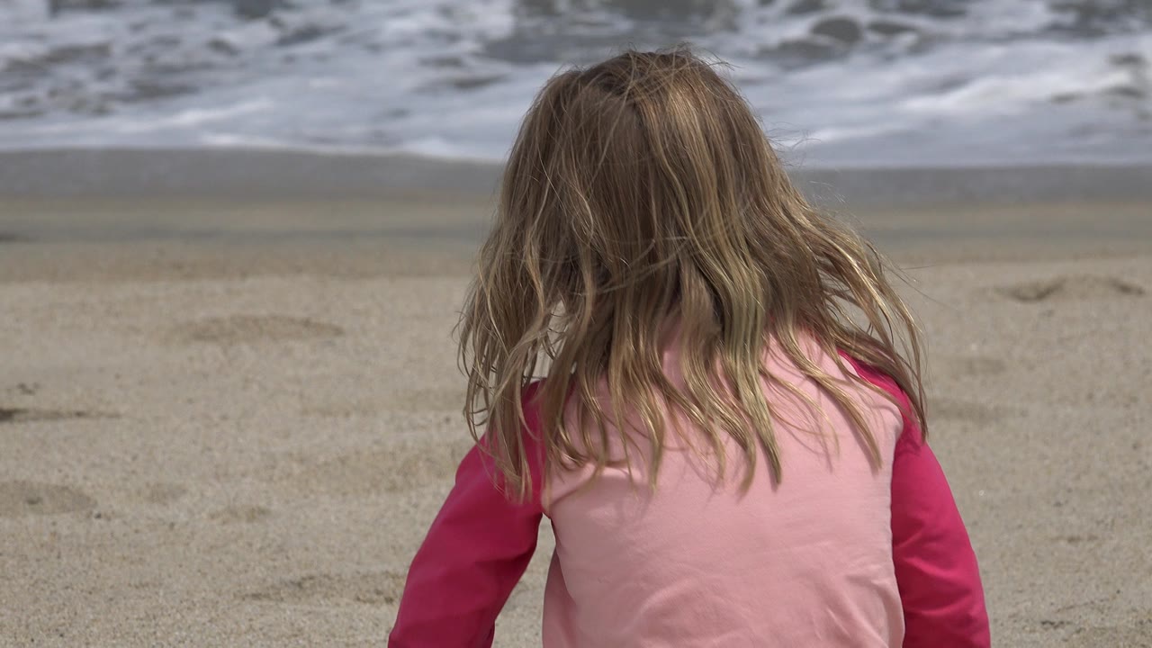 Young girl seen from behind sitting on sand near ocean foam.