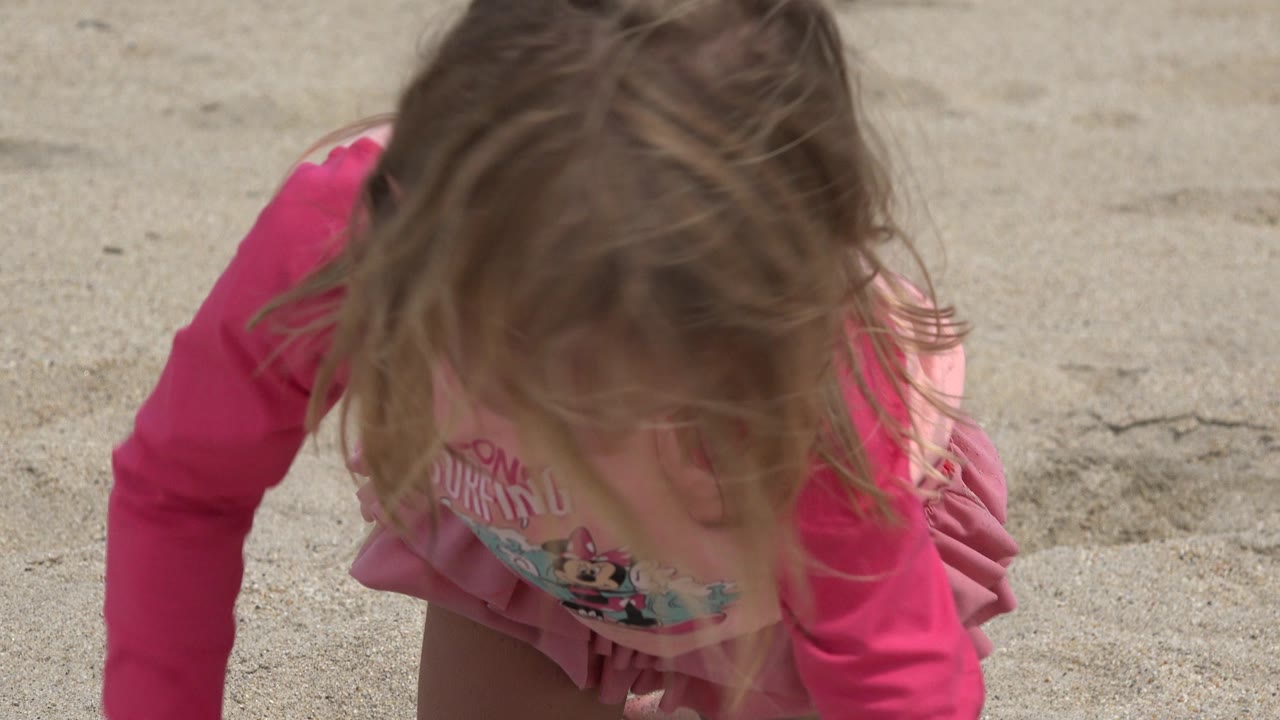 At 0:00, a young girl in a pink long-sleeve beach top leans forward over pale sand in San Jose del Cabo, Mexico, with her hair covering most of her face.