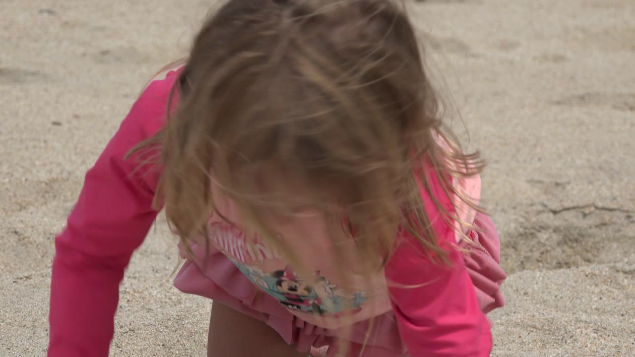 At 0:01, the child remains crouched close to the sandy beach in San Jose del Cabo, Mexico, with pink sleeves and windblown hair prominent in the frame.