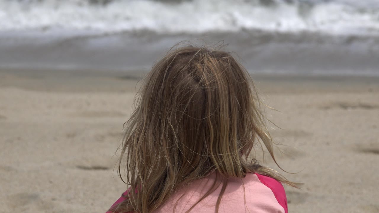 At 0:01, the child remains centered from behind on the San Jose del Cabo sand, with slightly shifting hair and pale surf spread across the background.