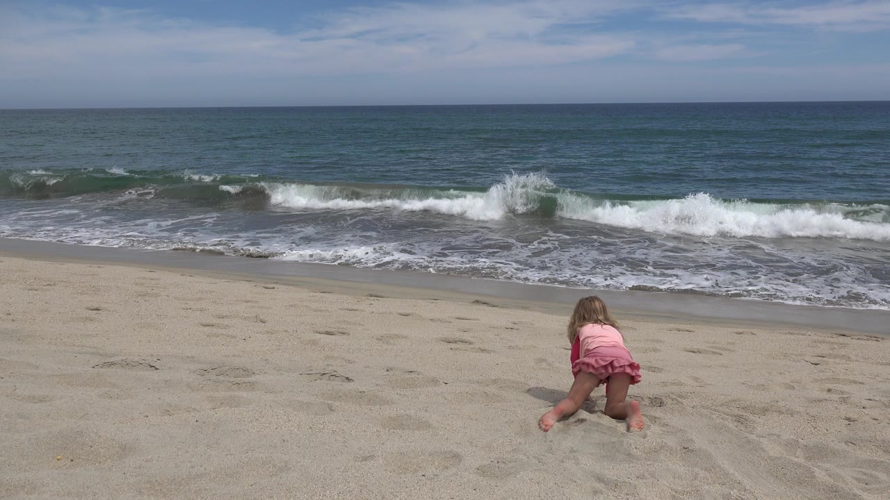 Young girl in pink on sand facing ocean waves.