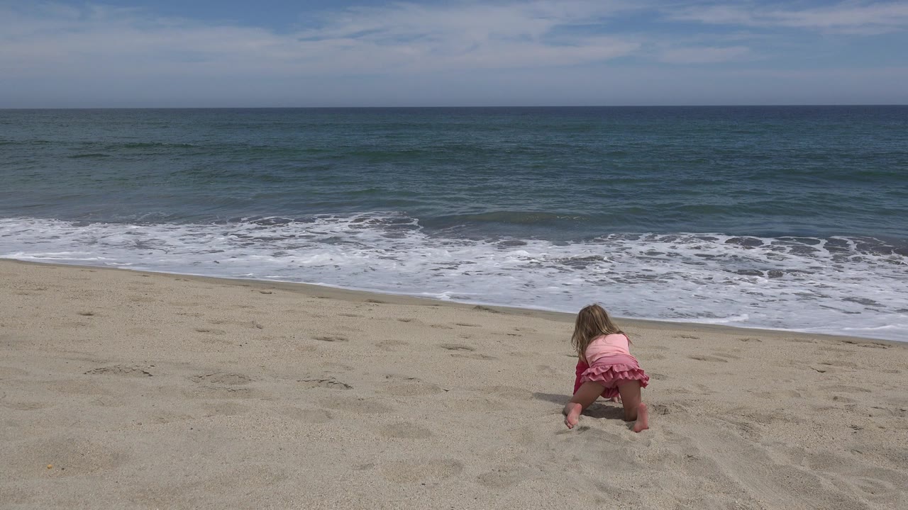 Young girl in pink crouching in beach sand near the ocean.
