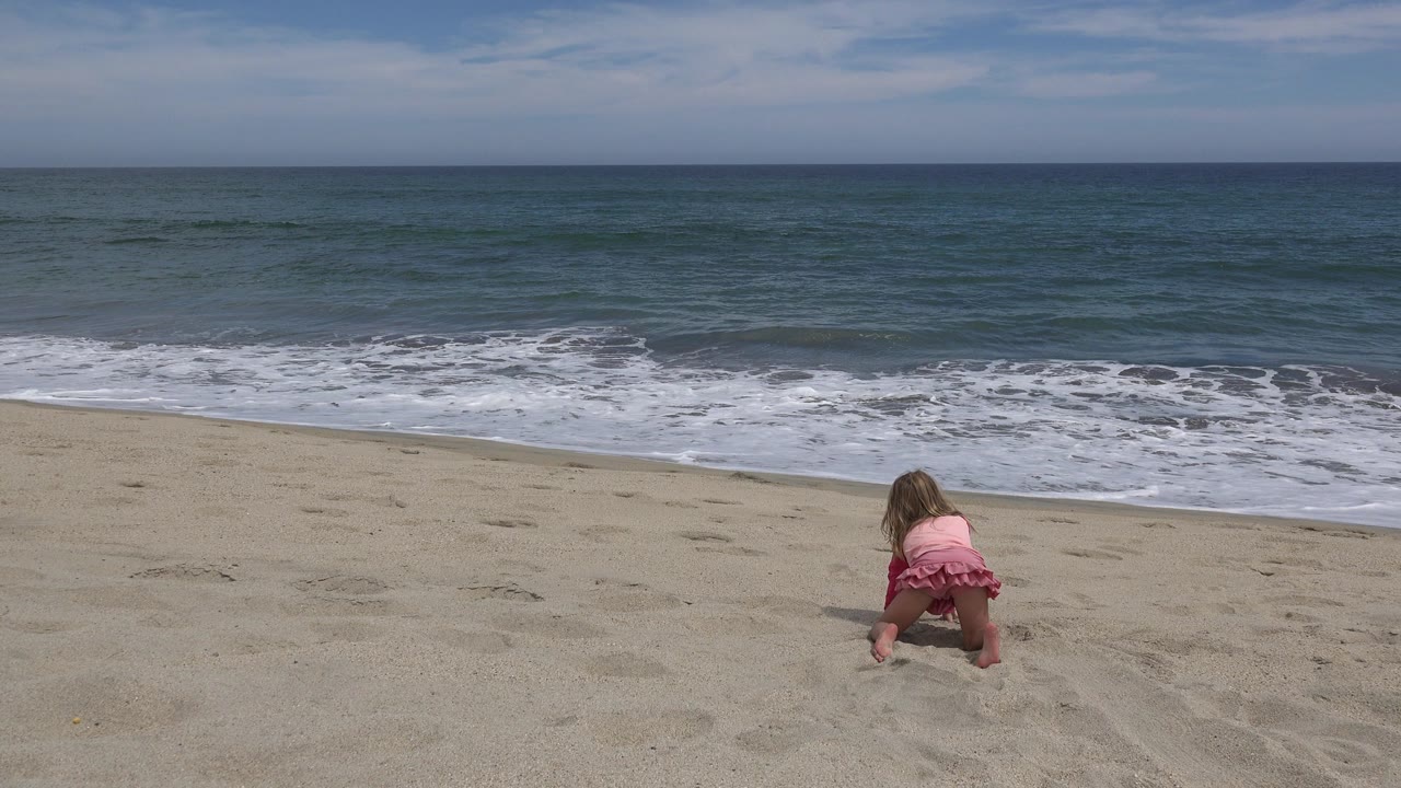 The girl remains kneeling near the center of the San Jose del Cabo beach frame as a band of foamy surf spreads along the shore behind her.