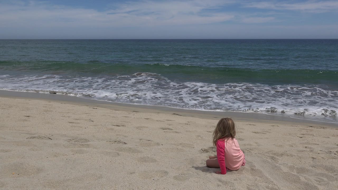 A child in a pink top sits near the lower center of a sandy beach in San Jose del Cabo, Mexico, facing small ocean waves under soft daylight.