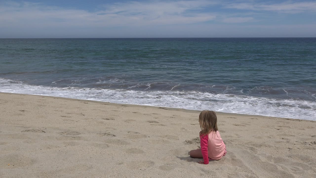 A young girl in pink sits on the sand facing the ocean in San Jose del Cabo, Mexico, with low surf and a broad horizon behind her.