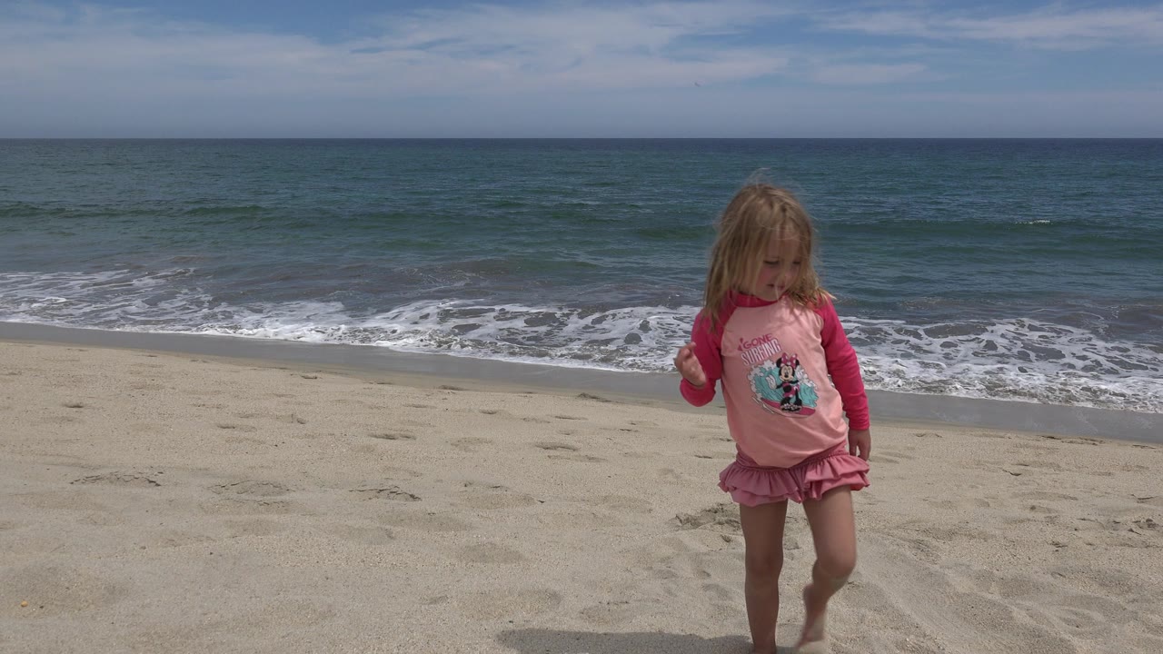 Young child walking on sand with ocean waves behind her.