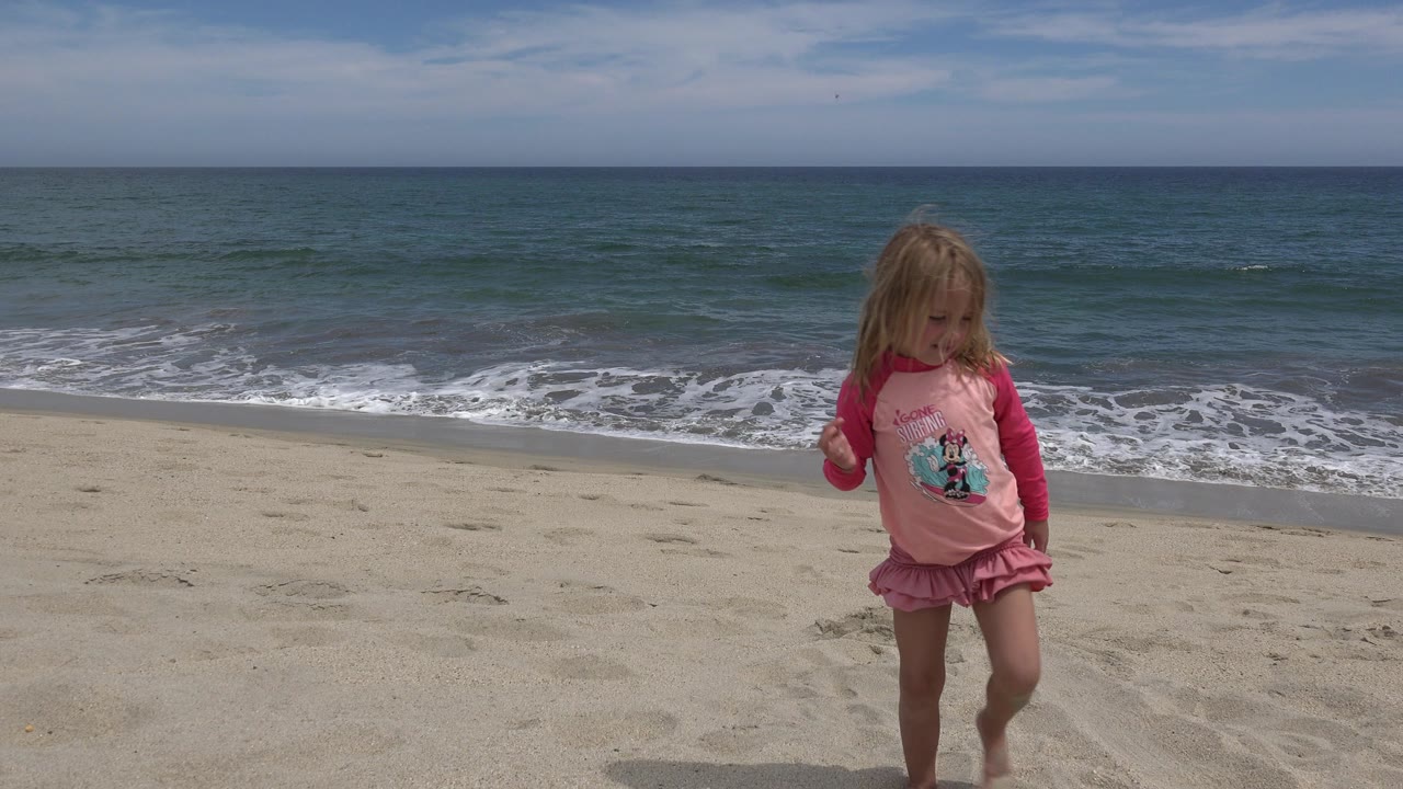The child continues walking across the San Jose del Cabo beach, with pale sand in the foreground and a thin band of breaking surf behind her.