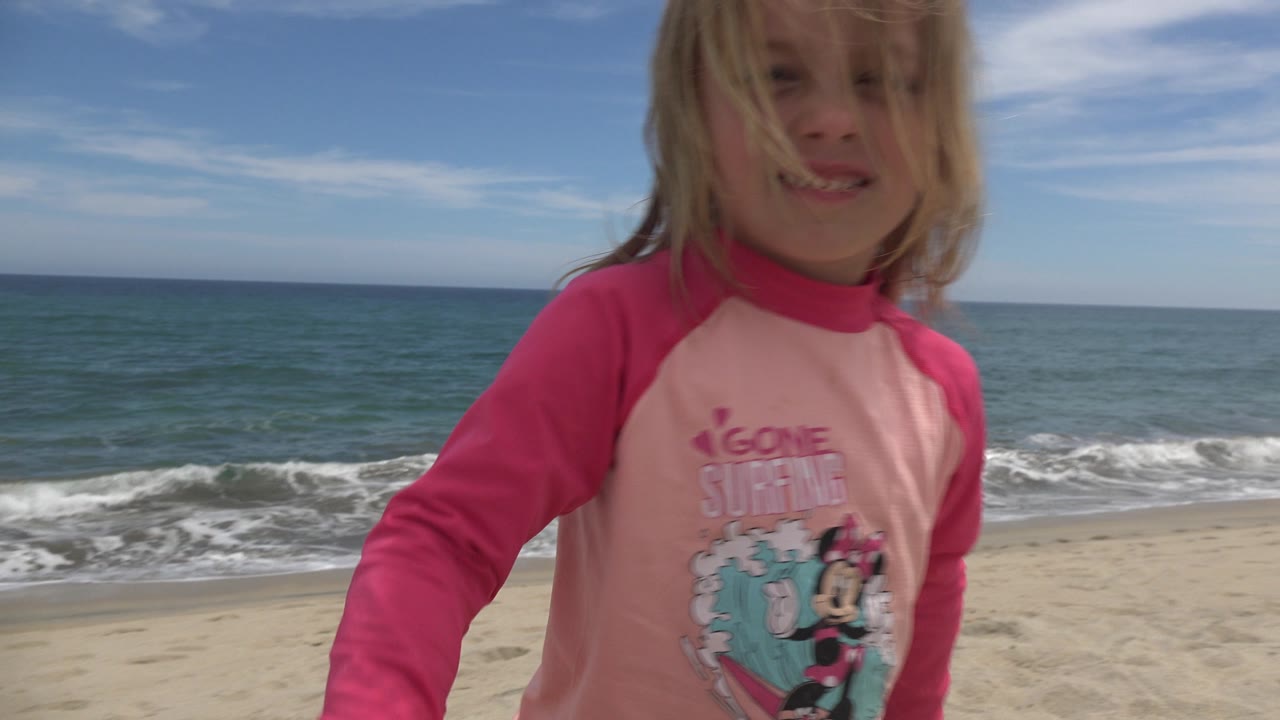The girl remains front-facing in San Jose del Cabo, with her hair across part of her face and small waves breaking along the sandy shoreline behind her.
