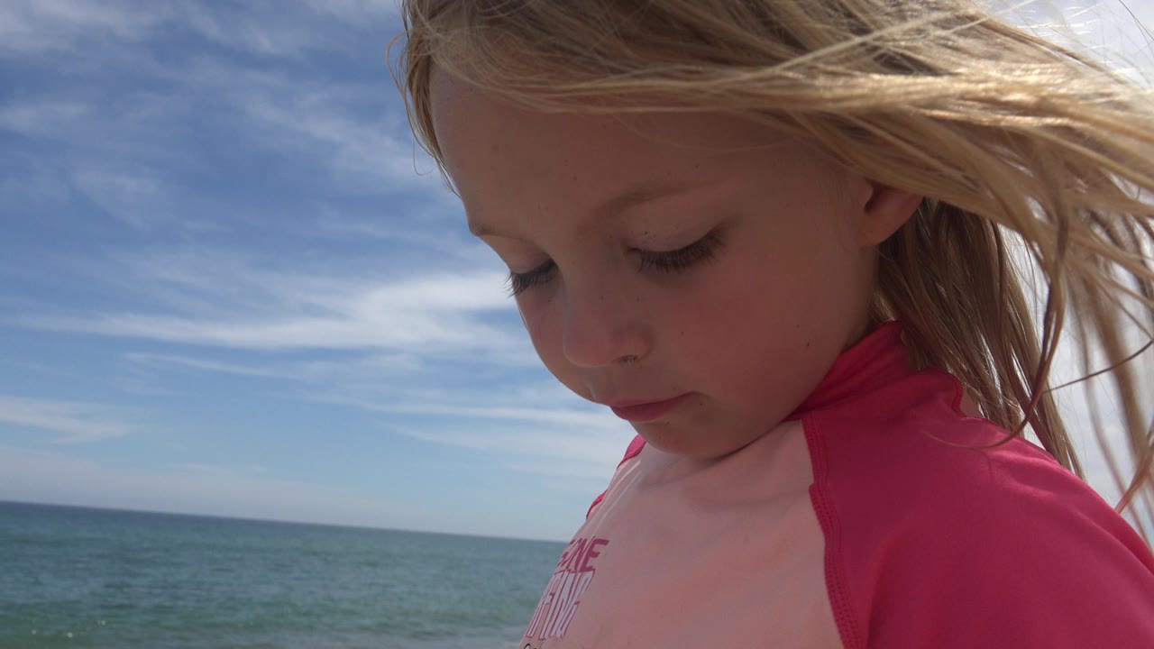 Close profile of a blonde child in a pink rash guard beside the ocean.