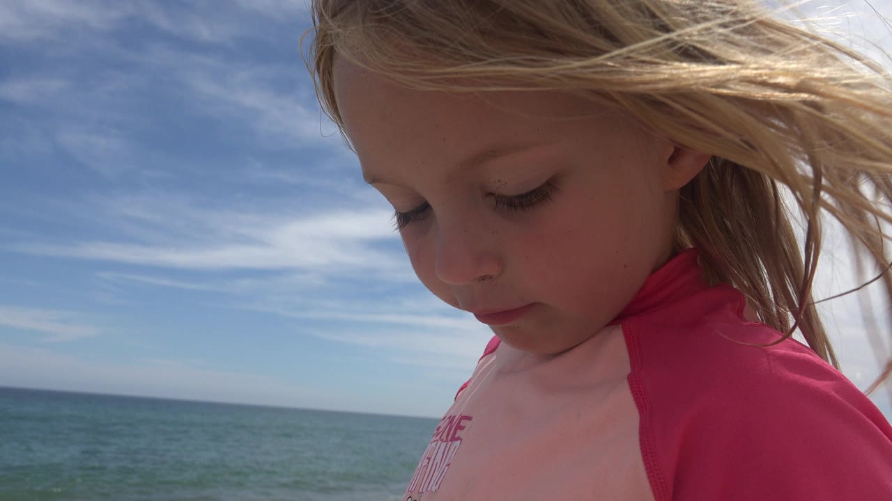 The child looks downward in a close San Jose Del Cabo beach frame, with windblown hair crossing the sky and the ocean horizon behind her.