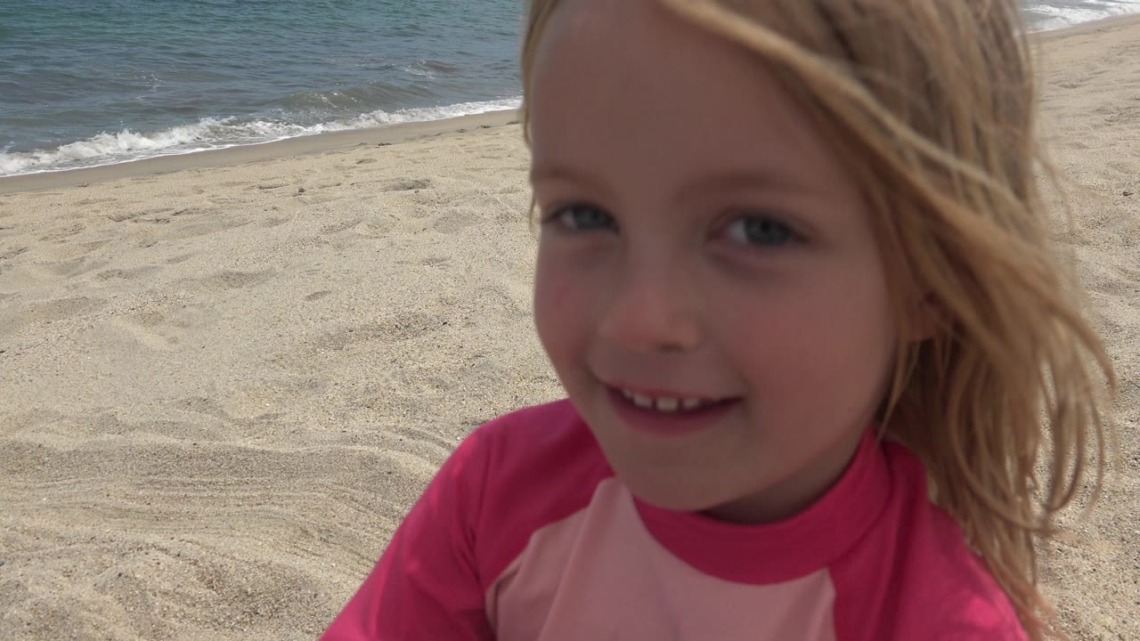 Young blonde girl close to camera on a sandy beach with ocean behind her.