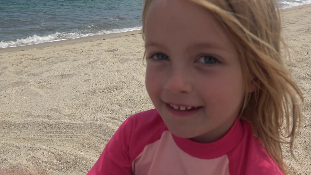 At 0:01, the blonde girl smiles near the lens in San Jose del Cabo, Mexico, while wind moves her hair and the beach slopes toward the ocean behind her.
