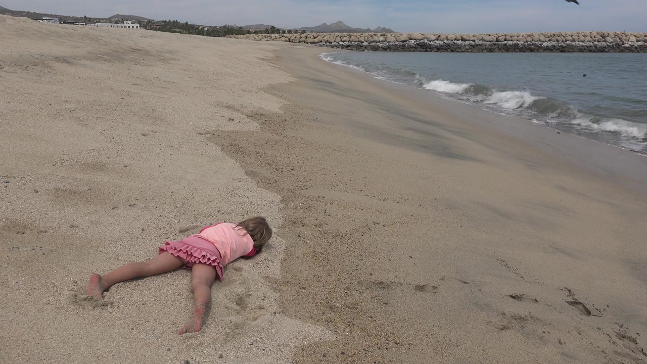 A child in pink remains on the sandy beach in San Jose del Cabo while a foamy wave line moves closer along the right edge of the shore.