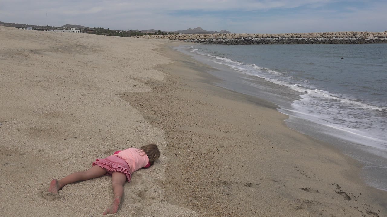 A young girl in pink lies face down on the sand near the shoreline in San Jose del Cabo, Mexico, with small waves and a rock breakwater behind her.