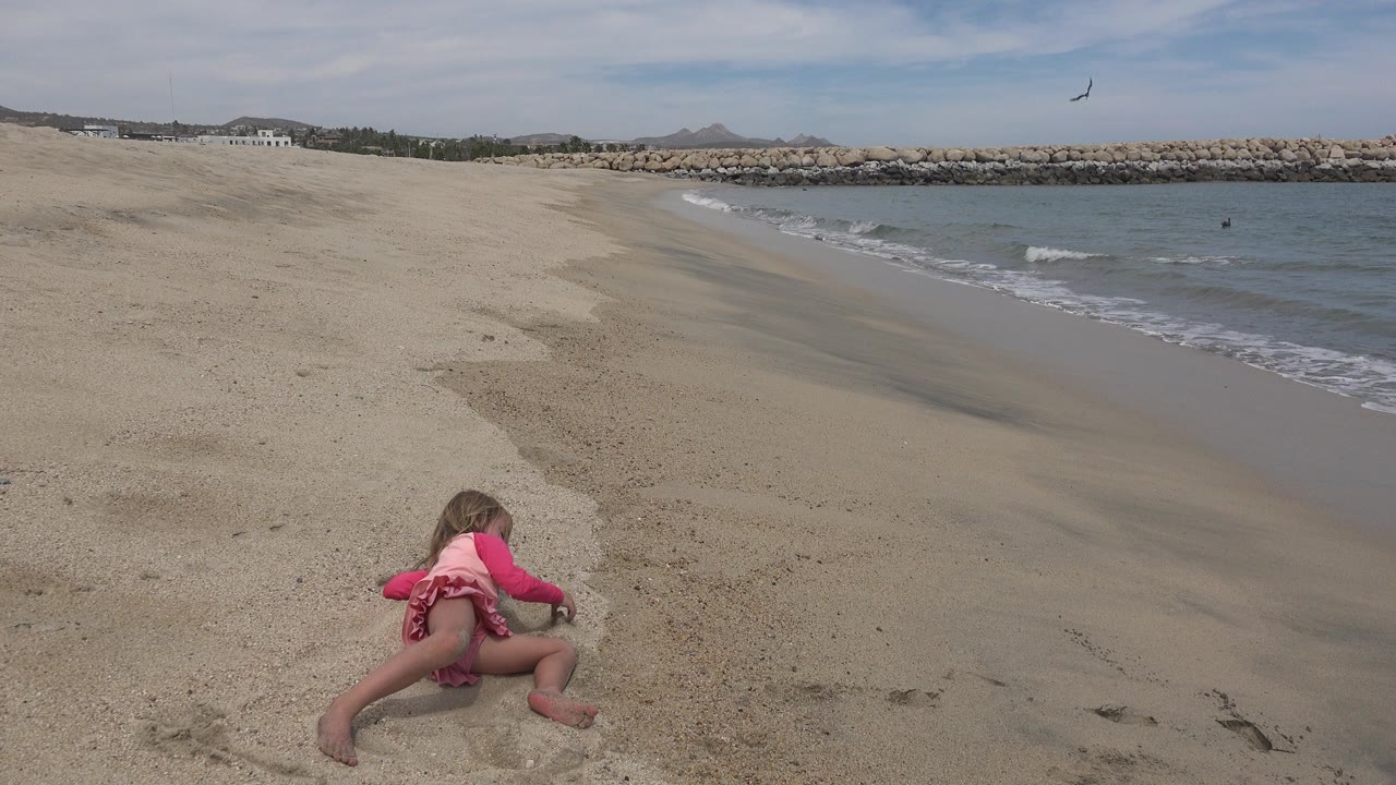A young child in pink clothing sits low on the sand near the ocean shoreline in San Jose del Cabo, Mexico, with small waves and a rock breakwater behind her.