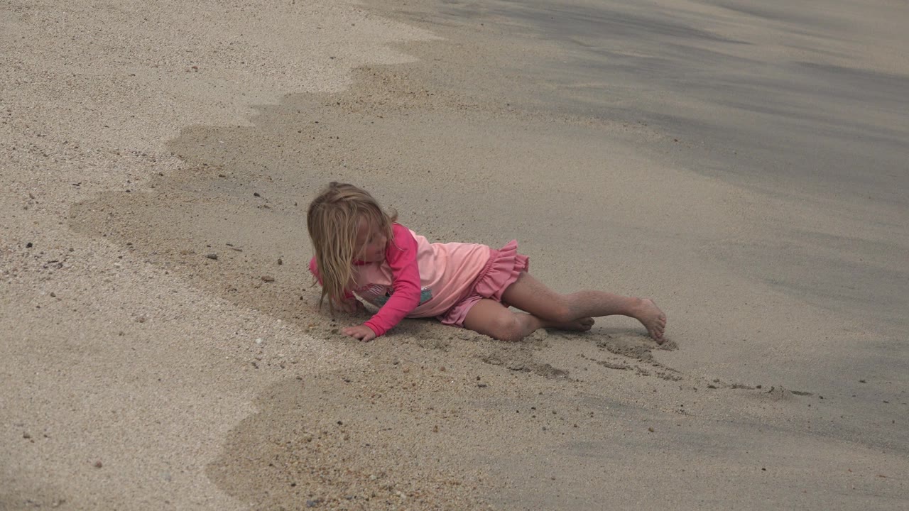 Young girl in a pink and red dress lying on sand near wet beach.